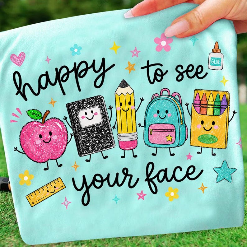 Puede incluir: Una camiseta azul claro con un gr&aacute;fico colorido de &uacute;tiles escolares con caras, incluyendo una manzana, un cuaderno, un l&aacute;piz, una mochila y crayones. El texto "happy to see your face" aparece en letra cursiva negra, rodeado de estrellas y flores.