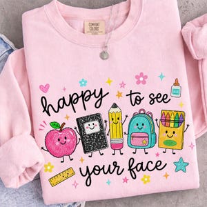 Puede incluir: Una sudadera de cuello redondo rosa claro con el texto "happy to see your face" e ilustraciones coloridas de dibujos animados de una manzana sonriente, un cuaderno, un l&aacute;piz, una mochila, una caja de ceras, pegamento y una regla.