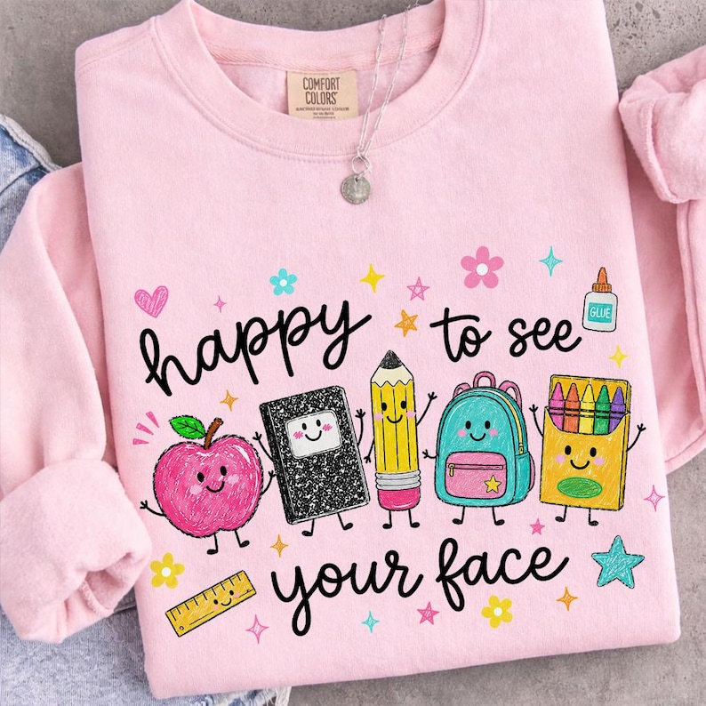 Puede incluir: Una sudadera de cuello redondo rosa claro con el texto "happy to see your face" e ilustraciones coloridas de dibujos animados de una manzana sonriente, un cuaderno, un l&aacute;piz, una mochila, una caja de ceras, pegamento y una regla.