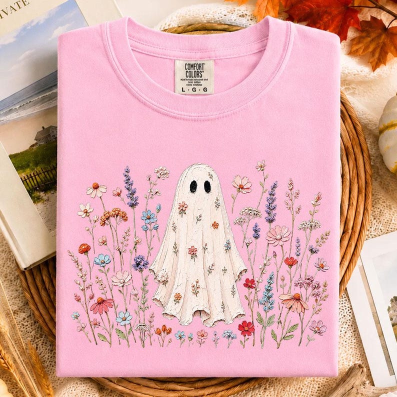 May include: A light pink crew neck t-shirt featuring a central graphic of a white ghost surrounded by a variety of colorful wildflowers in shades of blue, purple, orange, and pink. The interior neck label displays the text Comfort Colors L G G.
