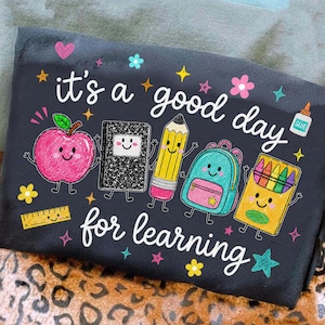 May include: A black t-shirt featuring the white text "it's a good day for learning" surrounded by colorful cartoon school supplies including a pink apple, a black notebook, a yellow pencil, a teal backpack, a crayon box, and a ruler.