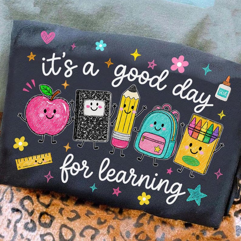 May include: A black t-shirt featuring the white text "it's a good day for learning" surrounded by colorful cartoon school supplies including a pink apple, a black notebook, a yellow pencil, a teal backpack, a crayon box, and a ruler.