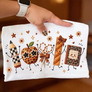 May include: A white t-shirt featuring an embroidered design of five anthropomorphic school supplies: a checkered pencil, a leopard-print apple with star-shaped sunglasses, scissors with a bow, a ruler with a bow, and a floral-patterned notebook.