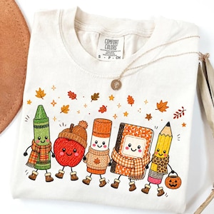 May include: A cream-colored t-shirt featuring a graphic of five anthropomorphic school supplies: a green crayon, a red apple, a glue stick, a notebook, and a yellow pencil. They wear autumn-themed scarves, hats, and boots, surrounded by falling leaves.