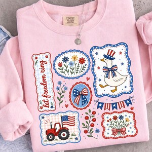 May include: A light pink sweatshirt with patriotic-themed graphics. The design includes a goose wearing a top hat and bow tie, a tractor, floral arrangements, and the text "let freedom ring."
