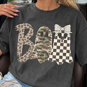 May include: A charcoal gray t-shirt featuring the word BOO in a graphic design. The letter B has a leopard print, the middle ghost has a green and brown camouflage pattern, and the final ghost has a black and white checkerboard pattern with a bow.