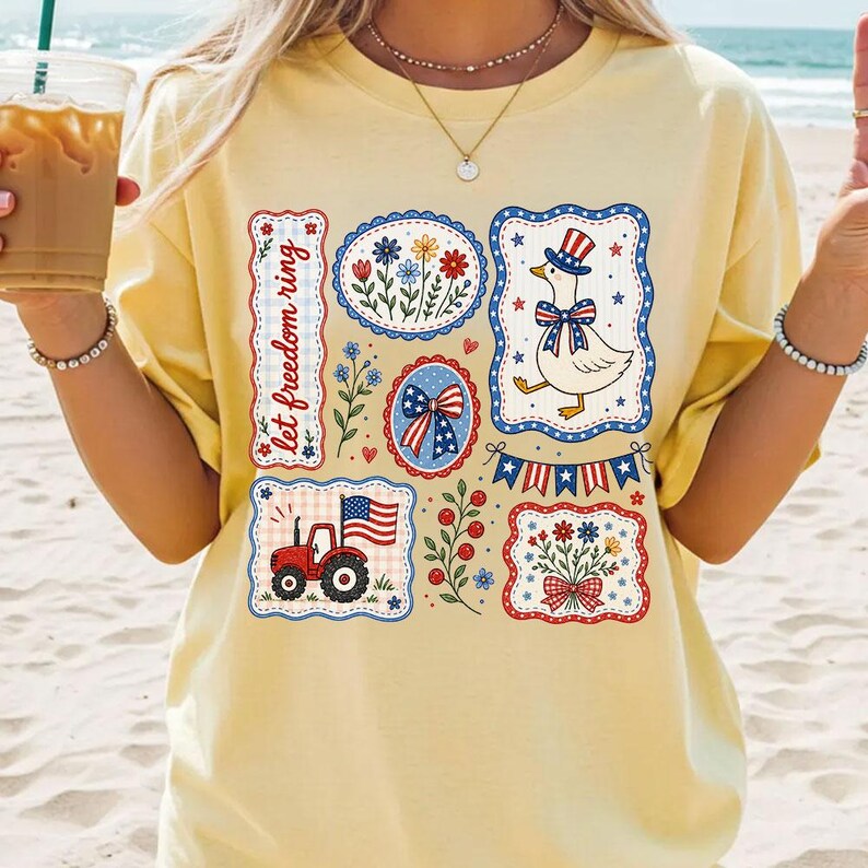 May include: A light yellow t-shirt featuring patriotic designs. The shirt displays various illustrations including a tractor, a goose wearing a hat, floral arrangements, and a bow, all framed in red, white, and blue. The text "let freedom ring" is also present.