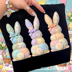 May include: Black tote bag featuring three appliqued Easter bunnies. Each bunny is made of patchwork fabric in pastel colors, with a bow and a rose at the base. The bunnies have floral and gingham patterns.