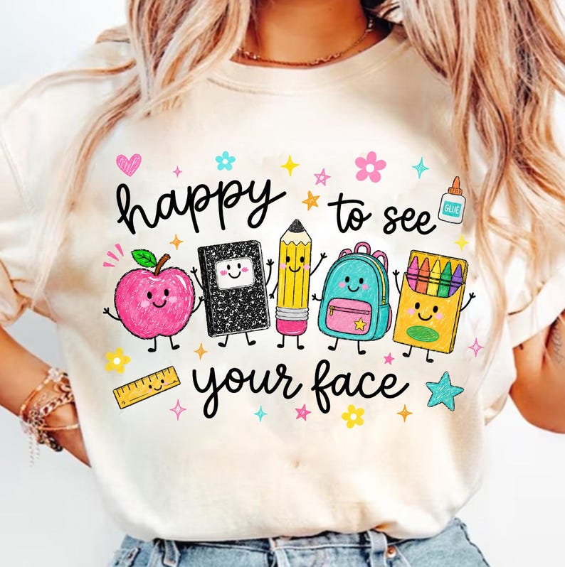 Puede incluir: Una camiseta de color crema con un gr&aacute;fico colorido y el texto "happy to see your face". El dise&ntilde;o incluye &uacute;tiles escolares estilo caricatura: una manzana rosa, un cuaderno negro, un l&aacute;piz amarillo, una mochila azul y una caja de crayones.