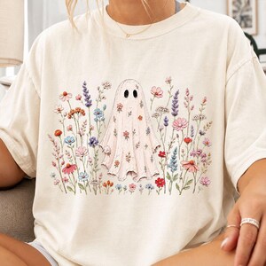May include: A cream-colored short-sleeve t-shirt featuring a central graphic of a ghost covered in small floral patterns, surrounded by a variety of colorful wildflowers including lavender, pink, blue, and orange blossoms across the front.