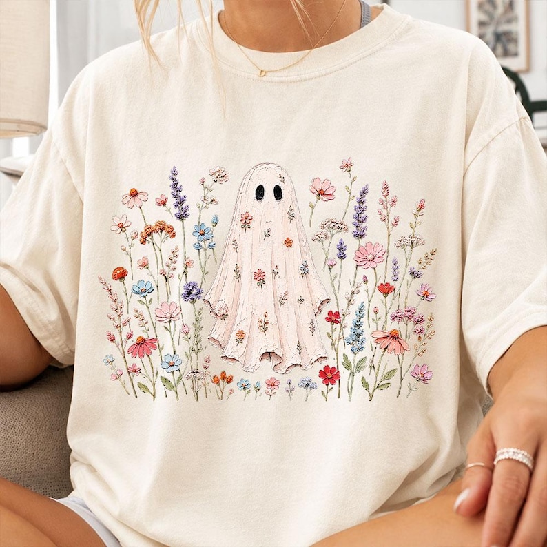 May include: A cream-colored short-sleeve t-shirt featuring a central graphic of a ghost covered in small floral patterns, surrounded by a variety of colorful wildflowers including lavender, pink, blue, and orange blossoms across the front.