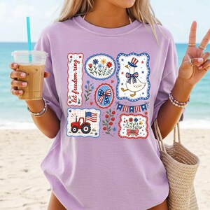 May include: A light purple t-shirt featuring patriotic designs. The shirt displays various patches with floral, flag, and tractor graphics. One patch reads "let freedom ring". The shirt is perfect for celebrating Independence Day.
