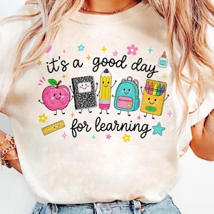 May include: A cream-colored t-shirt featuring a colorful graphic with the text 'it's a good day for learning'. The design includes cartoon illustrations of a pink apple, a black notebook, a yellow pencil, a blue backpack, and a box of crayons.