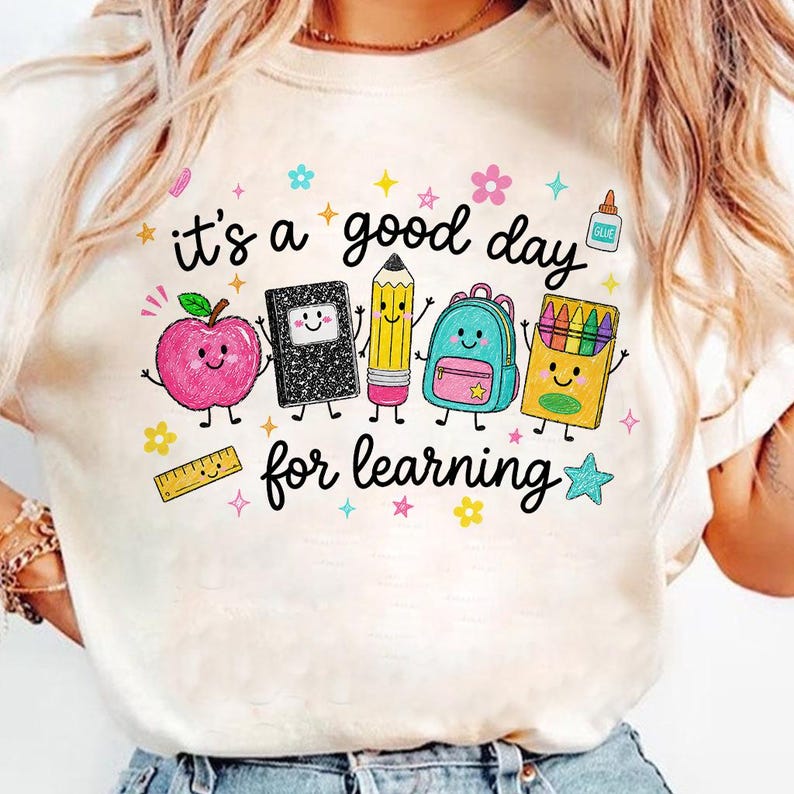 May include: A cream-colored t-shirt featuring a colorful graphic with the text 'it's a good day for learning'. The design includes cartoon illustrations of a pink apple, a black notebook, a yellow pencil, a blue backpack, and a box of crayons.