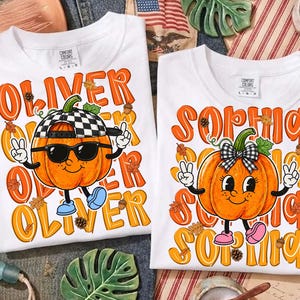 May include: Two white t-shirts featuring orange pumpkin graphics. The left shirt displays the name OLIVER with a pumpkin wearing a checkered cap and sunglasses. The right shirt displays the name SOPHIA with a pumpkin wearing a checkered bow.