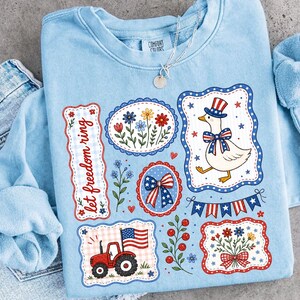 May include: Light blue sweatshirt with patriotic graphics. Features a cartoon goose in a top hat, a red tractor with an American flag, and floral designs. The text "let freedom ring" is also displayed.
