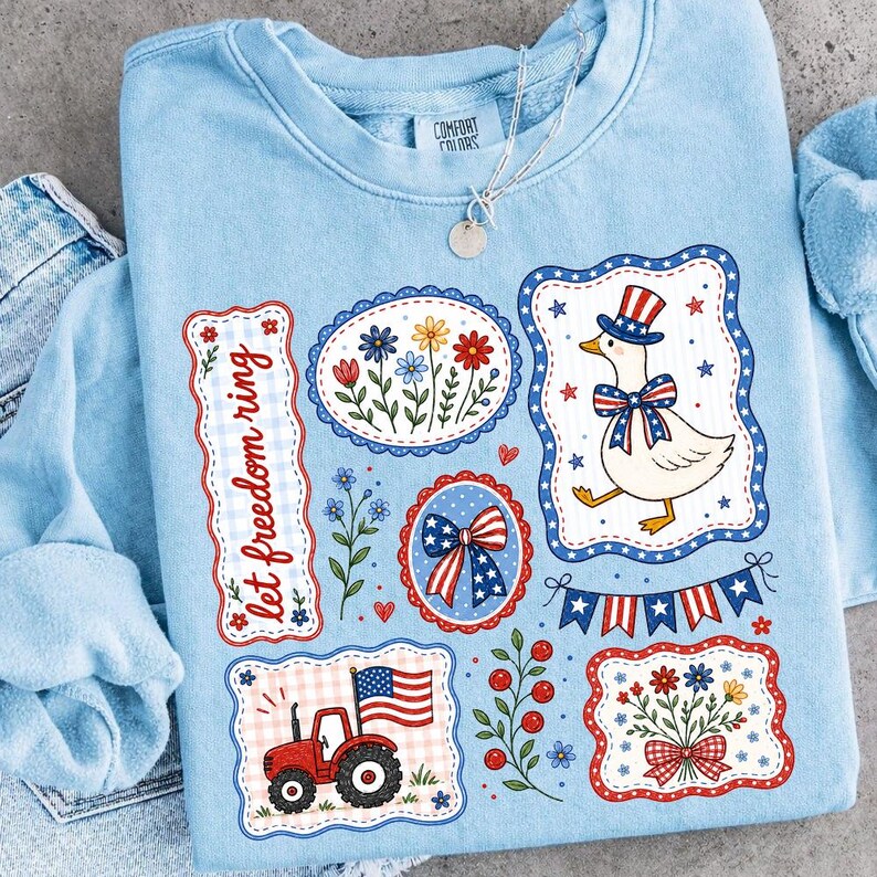May include: Light blue sweatshirt with patriotic graphics. Features a cartoon goose in a top hat, a red tractor with an American flag, and floral designs. The text "let freedom ring" is also displayed.