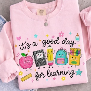 May include: A light pink crewneck sweatshirt featuring the text "it's a good day for learning" in black script. The graphic includes cartoon illustrations of a pink apple, a black composition notebook, a yellow pencil, a blue backpack, and crayons.