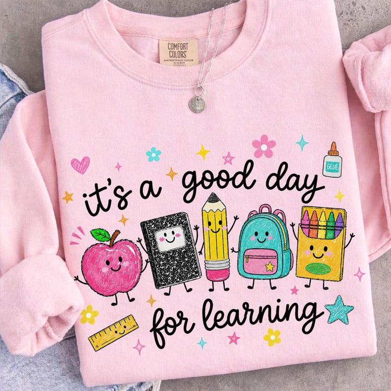 May include: A light pink crewneck sweatshirt featuring the text "it's a good day for learning" in black script. The graphic includes cartoon illustrations of a pink apple, a black composition notebook, a yellow pencil, a blue backpack, and crayons.
