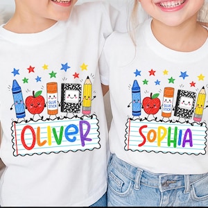 May include: Two white t-shirts featuring a colorful school-themed graphic. The design includes a cartoon crayon, apple, glue stick, notebook, and pencil above a lined paper rectangle with the names Oliver and Sophia printed in rainbow letters.