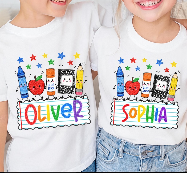 May include: Two white t-shirts featuring a colorful school-themed graphic. The design includes a cartoon crayon, apple, glue stick, notebook, and pencil above a lined paper rectangle with the names Oliver and Sophia printed in rainbow letters.