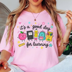 May include: A light pink t-shirt featuring a graphic print with the text "it's a good day for learning" surrounded by cartoon illustrations of a smiling apple, notebook, pencil, backpack, crayon box, glue bottle, ruler, and various colorful stars.