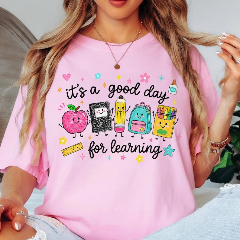 May include: A light pink t-shirt featuring a graphic print with the text "it's a good day for learning" surrounded by cartoon illustrations of a smiling apple, notebook, pencil, backpack, crayon box, glue bottle, ruler, and various colorful stars.