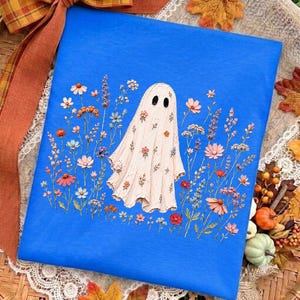 May include: A bright blue rectangular surface featuring a central illustration of a white ghost covered in small colorful flowers. The ghost is surrounded by a variety of delicate wildflowers in shades of pink, orange, blue, and purple.