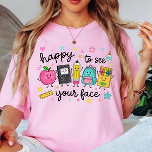 Puede incluir: Una camiseta rosa claro con un estampado gr&aacute;fico con el texto "happy to see your face" rodeado de coloridas e ilustraciones sonrientes de material escolar, incluyendo una manzana, un cuaderno, un l&aacute;piz, una mochila, ceras y una regla.