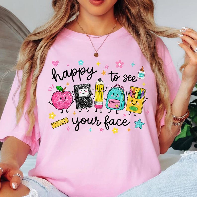 Puede incluir: Una camiseta rosa claro con un estampado gr&aacute;fico con el texto "happy to see your face" rodeado de coloridas e ilustraciones sonrientes de material escolar, incluyendo una manzana, un cuaderno, un l&aacute;piz, una mochila, ceras y una regla.