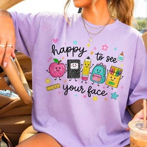 Puede incluir: Una camiseta de cuello redondo de color morado claro con un estampado gr&aacute;fico que incluye el texto "happy to see your face" rodeado de coloridas ilustraciones de material escolar sonriente, como una manzana, un cuaderno y un l&aacute;piz.