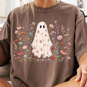 May include: A brown short-sleeve t-shirt featuring a central graphic of a white ghost surrounded by a variety of colorful wildflowers in shades of pink, blue, orange, and purple. The ghost figure is also decorated with small floral patterns.