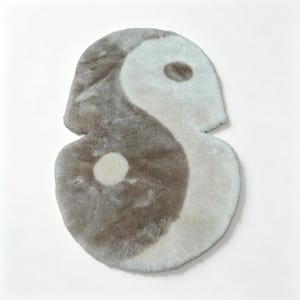 May include: A plush, faux fur rug shaped like a yin and yang symbol. The rug features a soft, shaggy texture in contrasting shades of cream and taupe, with a circular cream dot on the taupe side and a circular taupe dot on the cream side.