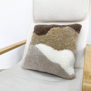 May include: A square decorative pillow with a textured, abstract design. The pillow features a pattern of brown, tan, white, and gray sections. It is placed on a white chair with wooden armrests, creating a cozy, neutral aesthetic.