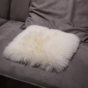 May include: A square, off-white sheepskin cushion rests on a gray sofa. The cushion has a fluffy, textured surface and appears soft. The sofa has a dark gray, velvety texture. The cushion is a decorative home accessory.