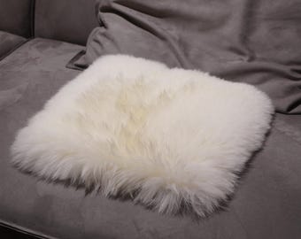 May include: A square, off-white sheepskin cushion rests on a gray sofa. The cushion has a fluffy, textured surface and appears soft. The sofa has a dark gray, velvety texture. The cushion is a decorative home accessory.