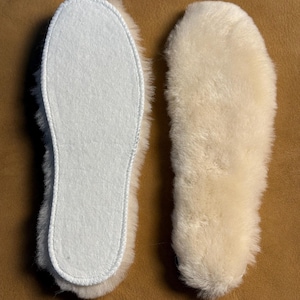 May include: Two sheepskin insoles. One insole is white with a stitched border and a white felt interior. The other insole is made of fluffy, cream-colored sheepskin. The insoles are designed to fit inside shoes.