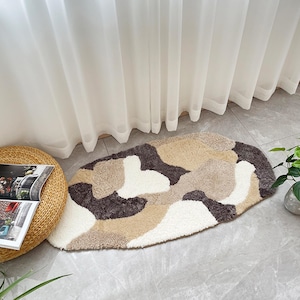 Pode incluir: Um tapete de banho oval com um padrão de camuflagem em tons de branco, bege e castanho. O tapete é feito de um material macio e felpudo e é colocado sobre um chão de azulejos perto de uma janela com cortinas transparentes. Uma banqueta de vime e uma planta também são visíveis.