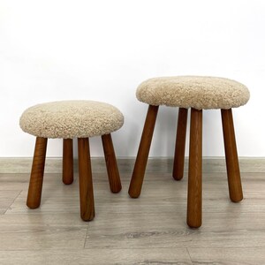 May include: Two fluffy, white, round, sheepskin covered stools with wooden legs. The stools are different sizes and are sitting on a light wood floor.