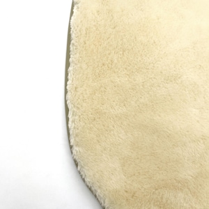 May include: Close-up of a cream-colored, oval-shaped rug with a plush, textured surface. The rug has a soft, fluffy appearance and a light brown border. The rug is on a white background.