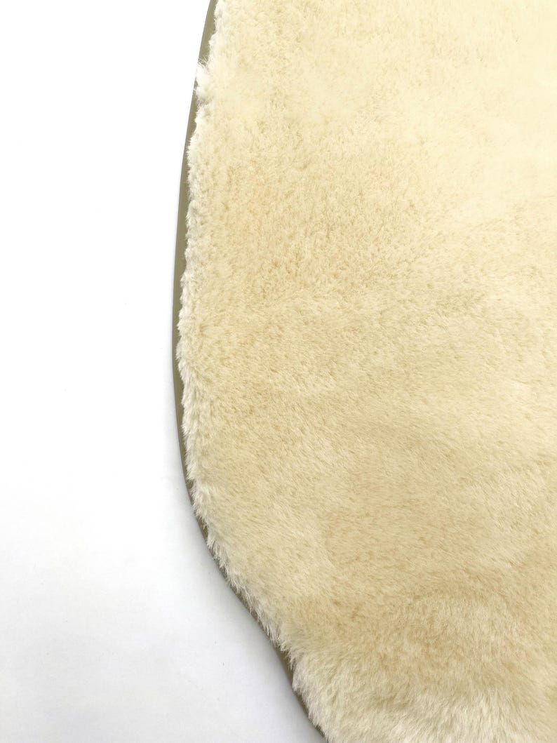 May include: Close-up of a cream-colored, oval-shaped rug with a plush, textured surface. The rug has a soft, fluffy appearance and a light brown border. The rug is on a white background.