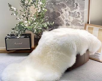 May include: A fluffy, off-white sheepskin rug draped over a brown leather ottoman. The rug has a soft, textured appearance. A Marshall speaker and framed artwork are in the background.