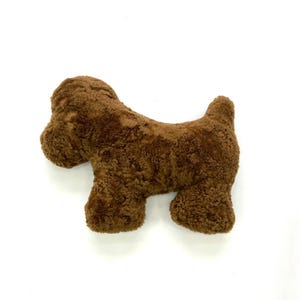 Puede incluir: Un cojín marrón con forma de perro. El cojín tiene una superficie texturizada y rizada y está diseñado en una forma simple y estilizada. El perro está representado de pie, con la cola levantada.