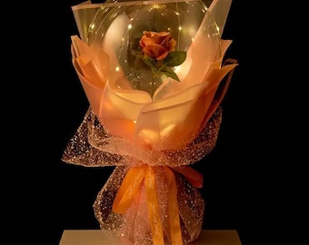 Peut inclure: Un bouquet de ballons transparent avec une seule rose orange et des lumières scintillantes à l'intérieur. Le bouquet est enveloppé de papier de soie rose et noué d'un ruban jaune.