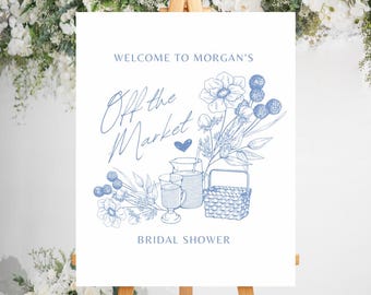 May include: A white sign with blue text and illustrations announcing a bridal shower. The text reads "Welcome to Morgan's Off the Market Bridal Shower". The illustration features a basket, pitchers, and flowers.