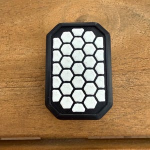May include: A rectangular black patch with a white honeycomb pattern. The patch has a black border and a geometric design. The object is set against a wooden background.