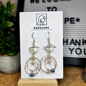 Puede incluir: Un par de pendientes dorados con un diseño de planeta. Los pendientes presentan un aro dorado, pequeñas perlas y una cuenta azul y morada. Los pendientes se exhiben en una tarjeta blanca con el texto "EARSCAPE YOUR SIGNATURE STATEMENT".