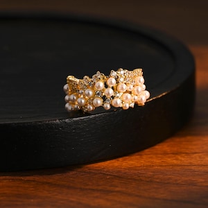 May include: A gold ring adorned with small pearls and clear crystals. The ring is set against a dark backdrop, accentuating its intricate design and elegant appearance. The ring is a band style, with multiple rows of pearls, creating a luxurious aesthetic.