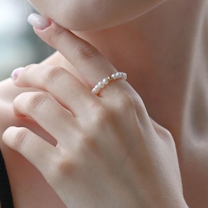 Könnte beinhalten: Ein zarter Ring mit einer Reihe kleiner, weißer Perlen. Ein einzelnes goldenes Kügelchen akzentuiert das Design. Der Ring wird an einem Finger getragen und zeigt seine elegante Schlichtheit und klassische Ausstrahlung. Die Perlen sind gleichmäßig angeordnet.