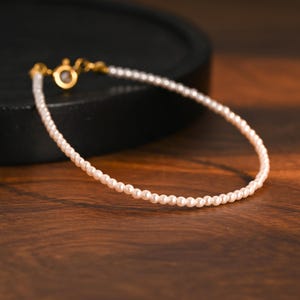 May include: A fine pearl bracelet with a gold clasp. The bracelet features small, round, white pearls. The background is a dark wooden surface with a black, circular object.